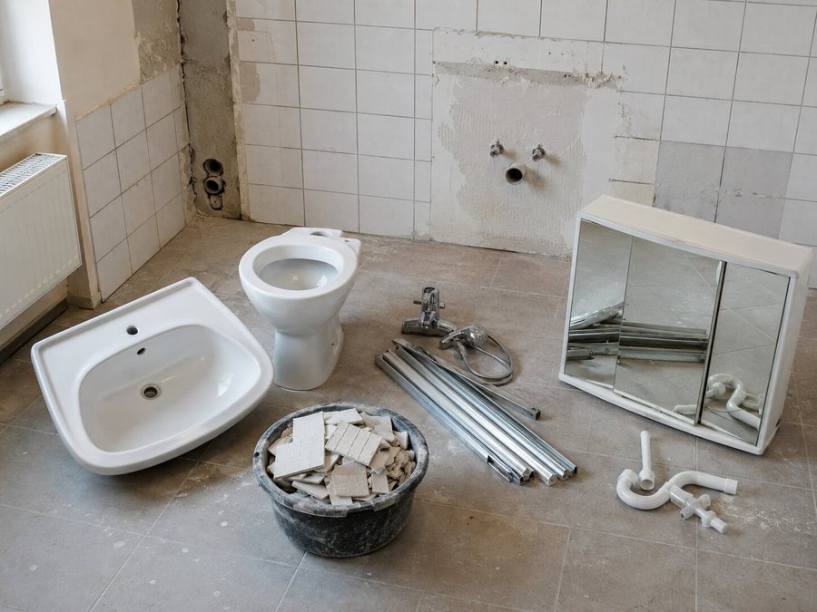 Teilweise zurückgebautes Badezimmer: ausgebautes Keramikwaschbecken und WC-Schüssel auf dem Boden, daneben ein Mörtelkübel mit Fliesenbruch, abmontierte Chromarmaturen und Aluminiumprofile, rechts ein abgenommener Spiegelschrank und weiße Kunststoff-Siphons.